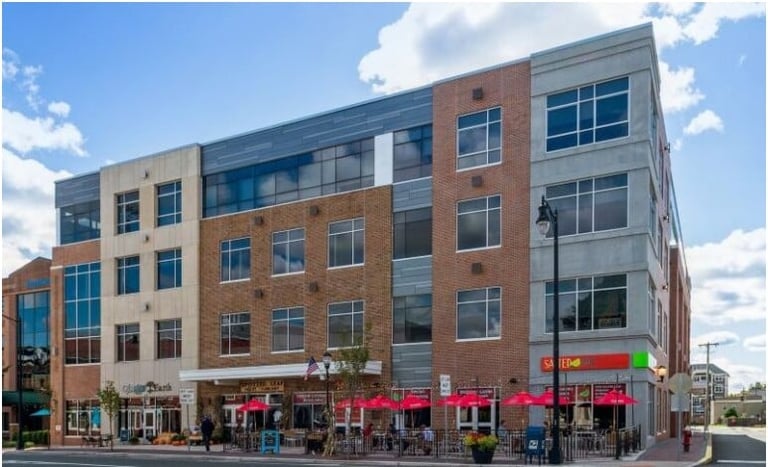 Modern mixed-use building with red brick and gray panels featuring street-level retail storefronts with colorful awnings and windows above.